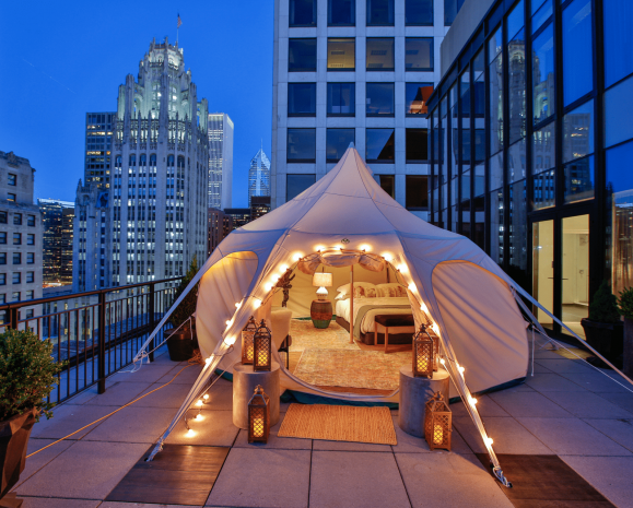 Tent with a bedroom set up on a downtown Chicago balcony with a cityscape in the background at nighttime.